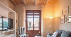 Un salon avec un téléviseur, un fauteuil à rayures, un canapé gris, une porte en bois donnant sur un beau paysage et une lampe fixée au mur.