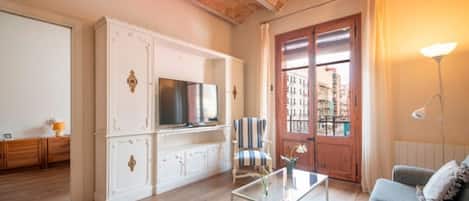 A living room with a sofa, a coffee table with magazines, a TV on a cabinet, and a view of buildings through a door.