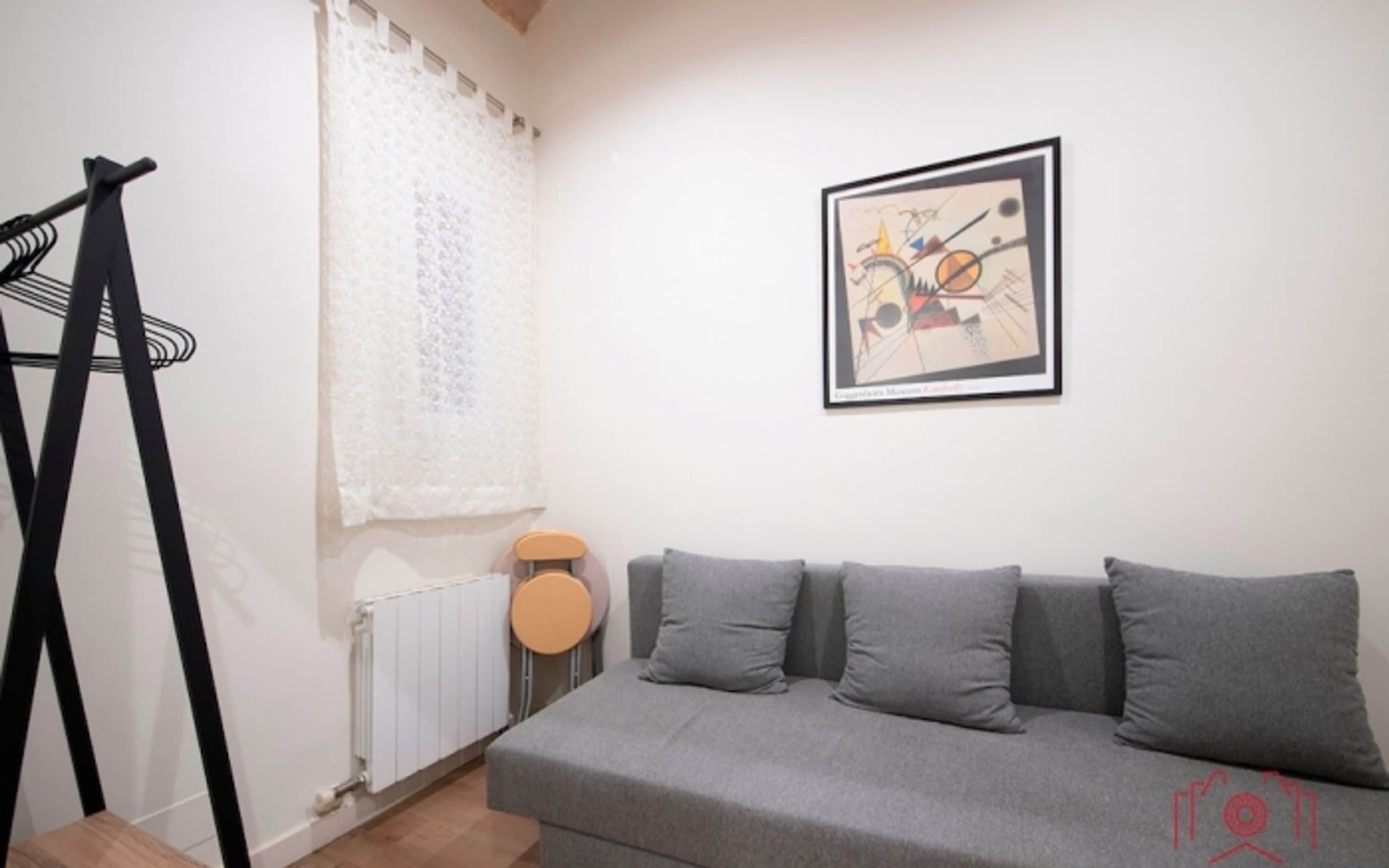 A modern living room with a grey sofa, a radiator, a window with white curtains, and a framed abstract painting on the wall.