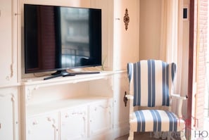 Ein Zimmer mit einem an einer weißen Kommode befestigten Fernseher, einem gestreiften Sessel und einem kleinen Tisch.