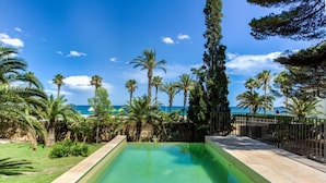A rectangular swimming pool with clear water, surrounded by a paved deck and a metal fence. Palm trees and other greenery are visible around the pool area. - Th06 Villa in Miami Platja 6 Bedrooms (Mont-roig del Camp)