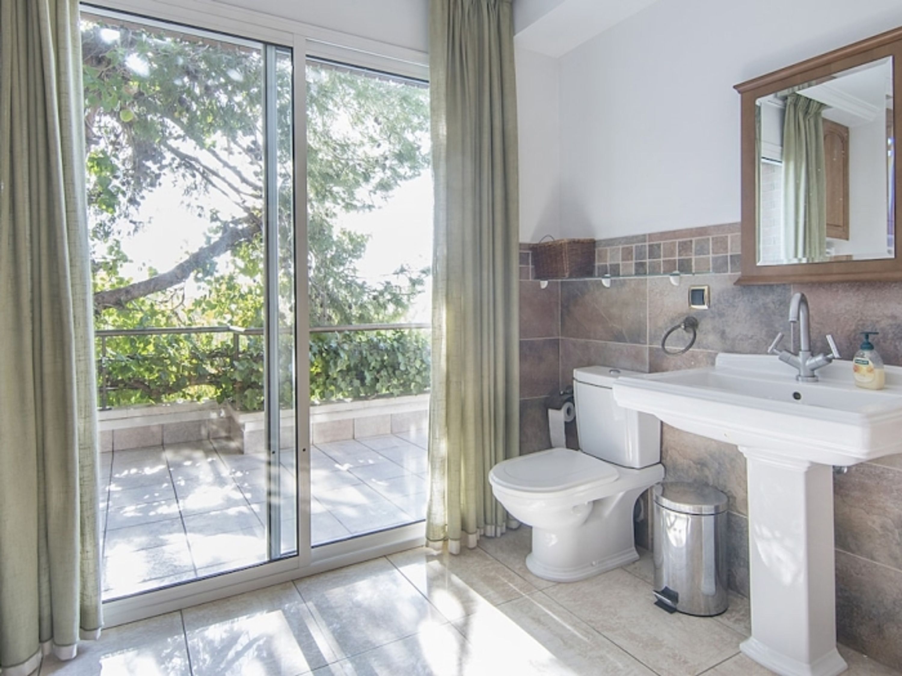 A bathroom with a toilet, sink, and trash can, a large window with curtains, and a mirror above the sink.