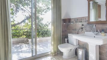 A bathroom with a toilet, sink, and trash can, a large window with curtains, and a mirror above the sink.
