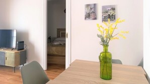 Une pièce avec une table en bois, un vase vert contenant des fleurs jaunes, un lit à l’arrière-plan et deux tableaux encadrés au mur.