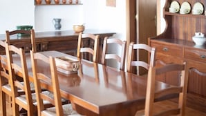 A wooden dining table with chairs, a wooden cabinet with shelves, and a kitchen area with pots and a kettle.