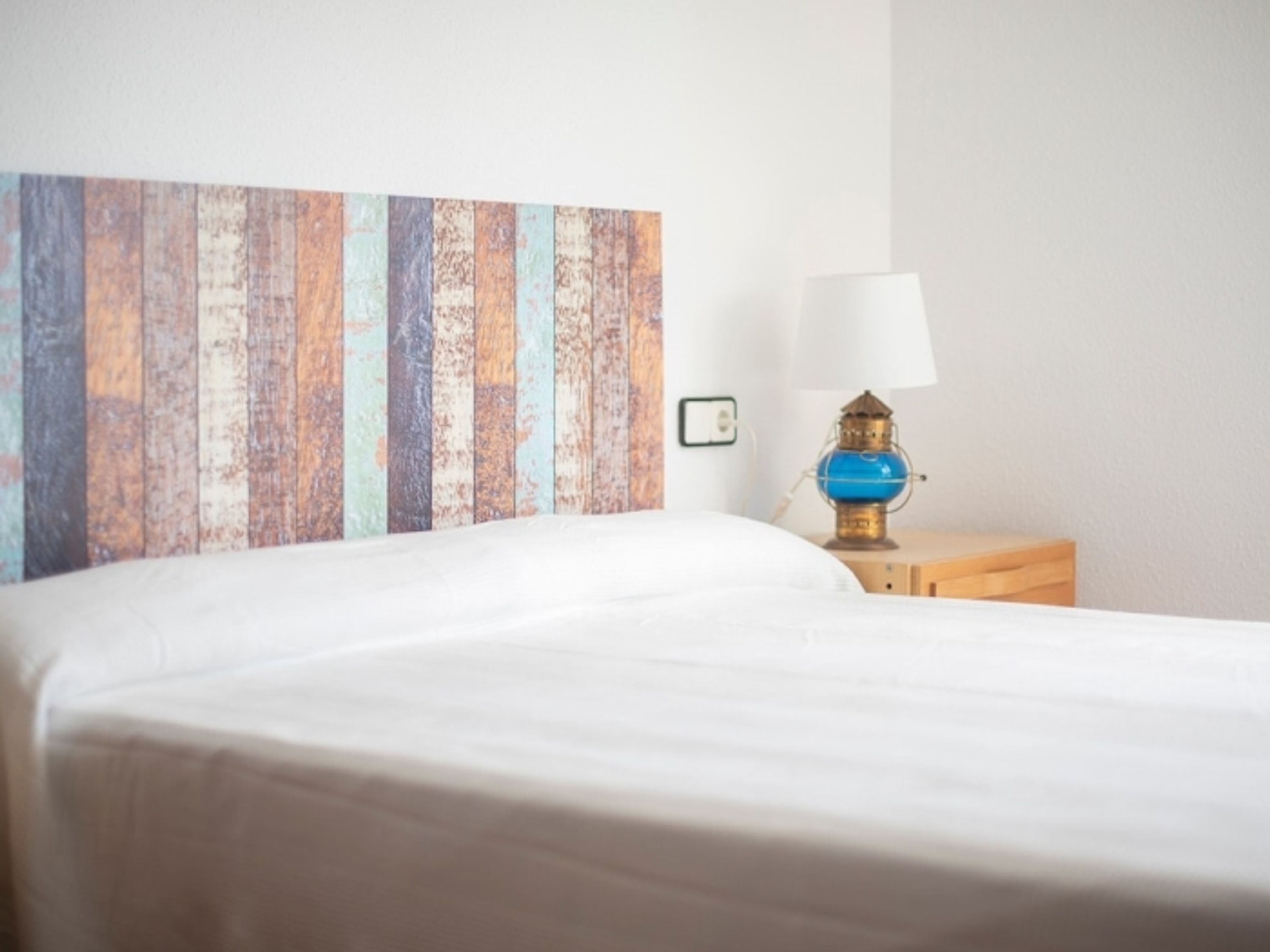 A neatly made bed with a white sheet, a wooden nightstand with a blue lamp, and a colorful wooden headboard.