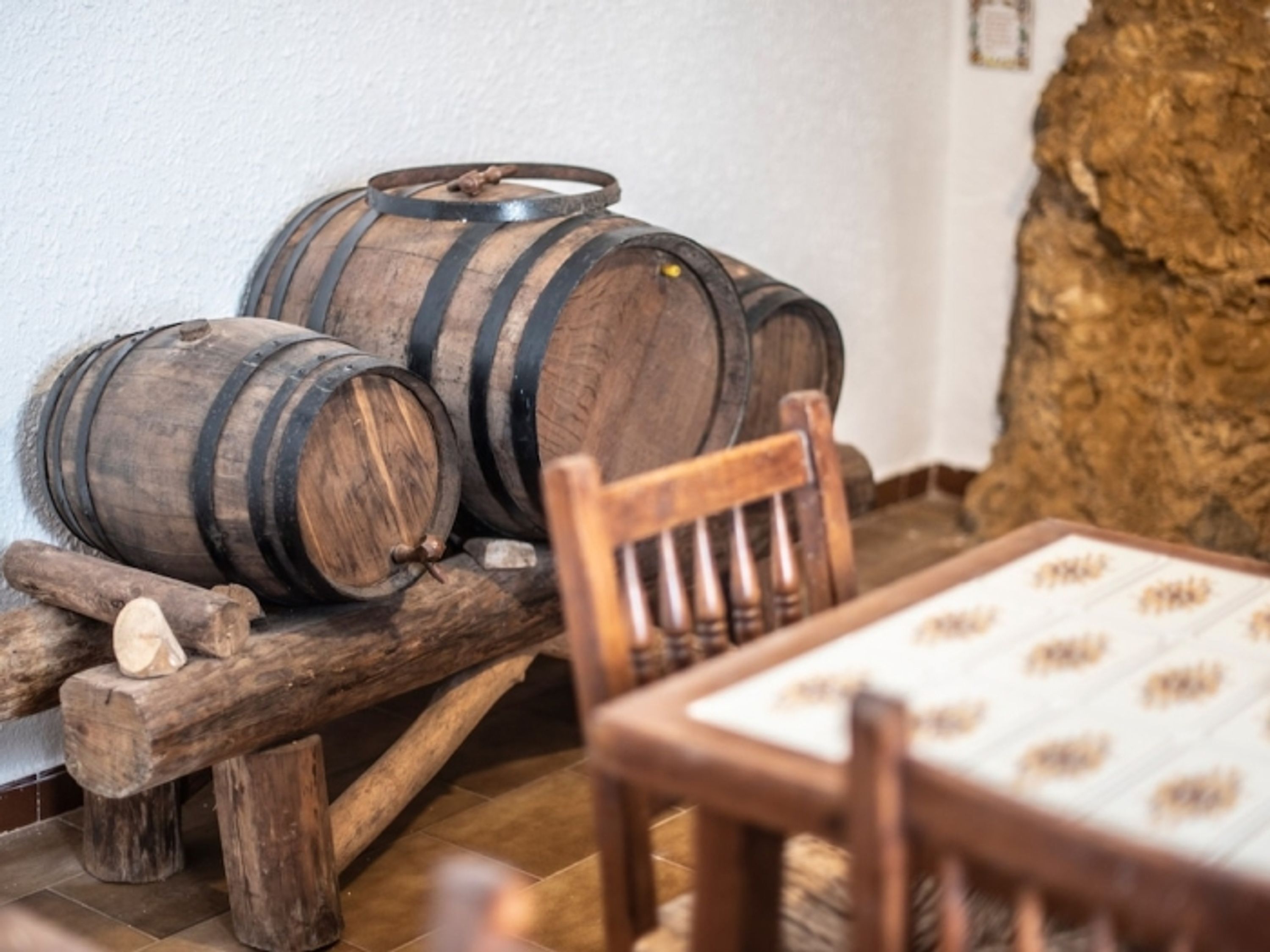 Barricas de madera apiladas sobre un banco de madera, una silla de madera rústica y una mesa con mantel estampado.
