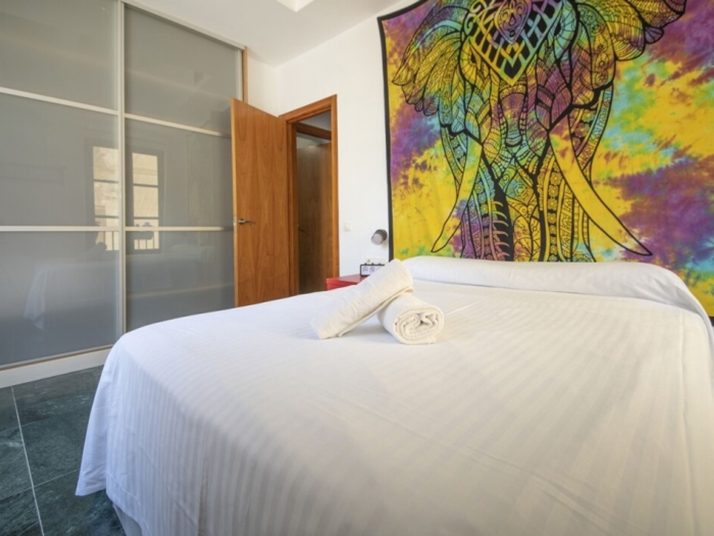 A hotel room with a bed, a large colorful tapestry, a wooden door, and a sliding glass door.