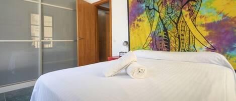 A hotel room with a bed, a large colorful tapestry, a wooden door, and a sliding glass door.