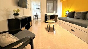 Un salon moderne avec un canapé, un téléviseur fixé au mur, un coin repas avec une table et des chaises, et une cuisine en arrière-plan.