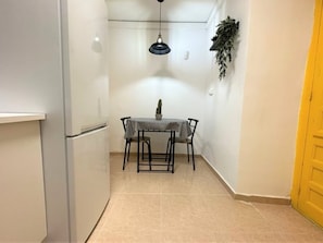 A compact kitchen with a refrigerator, a dining table with chairs, a hanging light fixture, and a potted plant.