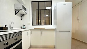 Une cuisine avec des meubles blancs, un four à micro-ondes, un réfrigérateur et une fenêtre avec des stores.