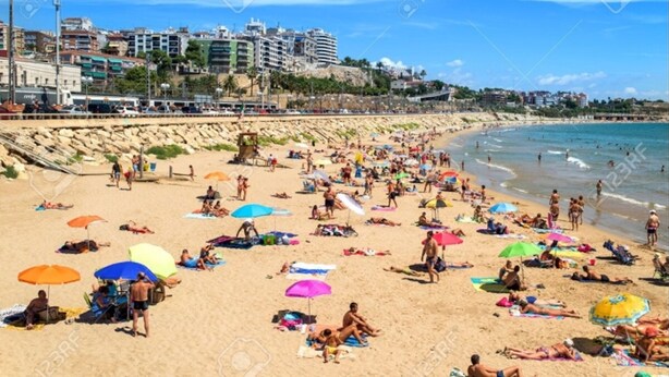 Una playa concurrida, gente tomando el sol y nadando, una costa rocosa y edificios al fondo.