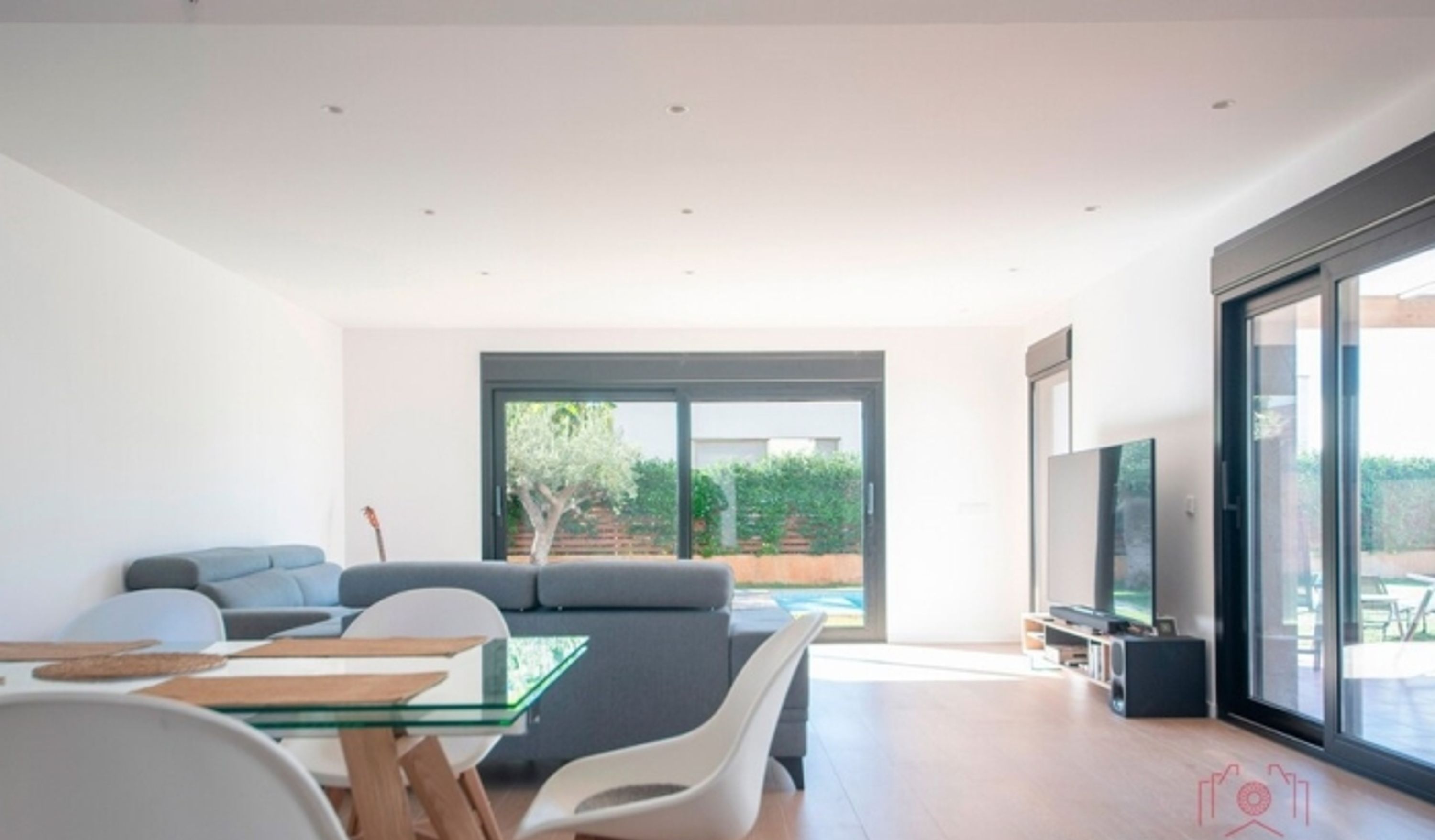 A modern living room with a glass dining table, white chairs, a grey sofa, and a TV on a stand.