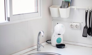 Eine moderne Küche mit Spüle aus Edelstahl, einer weißen Arbeitsplatte, einer Kaffeemaschine und einer Reihe schwarzer Küchenutensilien, die an der Wand hängen.