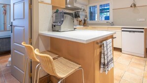 Una cocina compacta con una isla central, estufa integrada y lavavajillas. Hay dos sillas de mimbre y una toalla colgada en la isla.