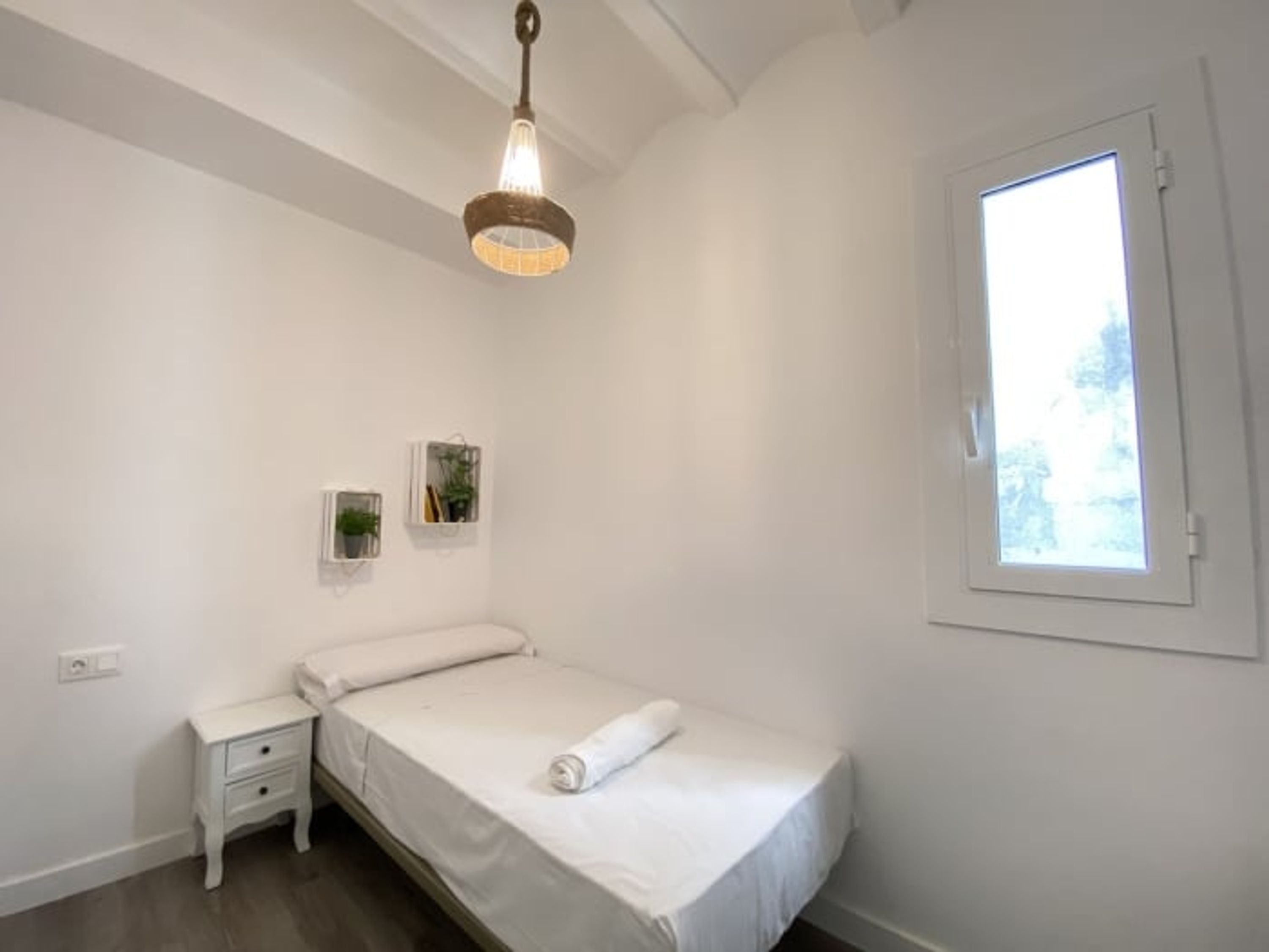 Ett litet, städat sovrum med en enkelsäng, ett sängbord, en hängande lampa, ett fönster och två växter som hänger på väggen.