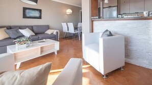 A modern living room with a grey sofa, white armchairs, and a coffee table. There is a dining area with white chairs and a kitchen with white cabinets. - Th123 Apartamento con Vistas al mar Cala Romana (Tarragona)
