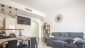 A modern living room with a grey sofa, a wooden dining table with chairs, and a kitchen area with a fridge and a TV. - Th134 Apartamento Moderno Centro de Tarragona (Tarragona)