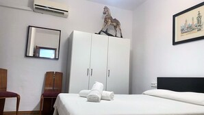 Una habitación de hotel con cama, armario, silla, unidad de aire acondicionado, espejo y un cuadro en la pared.