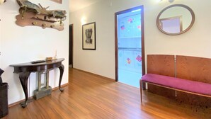 Una habitación con suelo de madera, un piano, un sofá, un espejo y una puerta que da a una cocina.