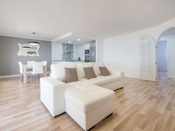 A modern living room with a white sectional sofa, a dining area with a white table and chairs, and a kitchen in the background. - Th88 Apartamento en Miami Playa (Mont-roig del Camp)