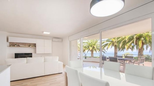 A modern living room with a white sofa, a TV, and a dining area with white chairs. There is a large window with a view of palm trees and the sea. - Th88 Apartamento en Miami Playa (Mont-roig del Camp)