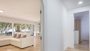 A modern living room with a white sofa, wooden flooring, and large windows overlooking palm trees. - Th88 Apartamento en Miami Playa (Mont-roig del Camp)