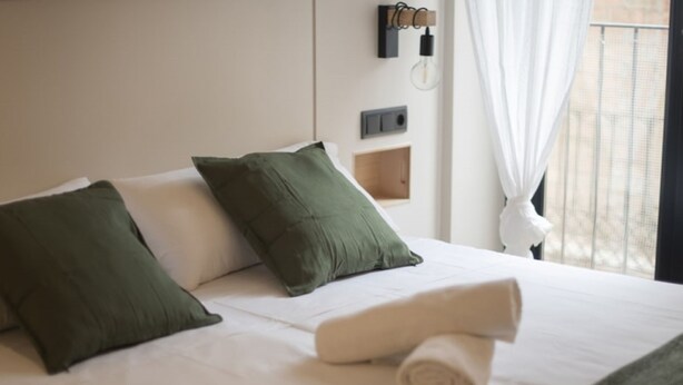 A neatly made bed with green and white pillows, a rolled towel, and a wall-mounted light fixture. - Th86 Apartamento con Encanto en Parte Alta (Tarragona)