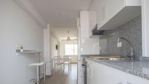 A modern kitchen with white cabinets, a granite countertop, a stainless steel sink, and a dining area with a chair and table. - Th64 Apartamento Moderno Cerca de la Universidad (Tarragona)