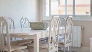 Ein Essbereich mit weißen Stühlen und einem Tisch, ein gewebter Korb auf dem Tisch und ein Fenster mit Blick auf Gebäude.