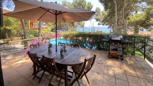 A patio with a round wooden table and chairs, a barbecue grill, and a view of the sea. - Th100 Villa en Tamarit con Vistas al mar (Tarragona)