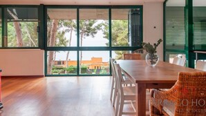 A spacious room with a wooden floor, a dining table with chairs, and large windows offering a view of trees and buildings outside. - Th100 Villa en Tamarit con Vistas al mar (Tarragona)