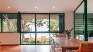 A spacious room with a wooden floor, a dining table with chairs, and large windows offering a view of trees and buildings outside.
