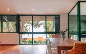 A spacious room with a wooden floor, a dining table with chairs, and large windows offering a view of trees and buildings outside. - Th100 Villa en Tamarit con Vistas al mar (Tarragona)