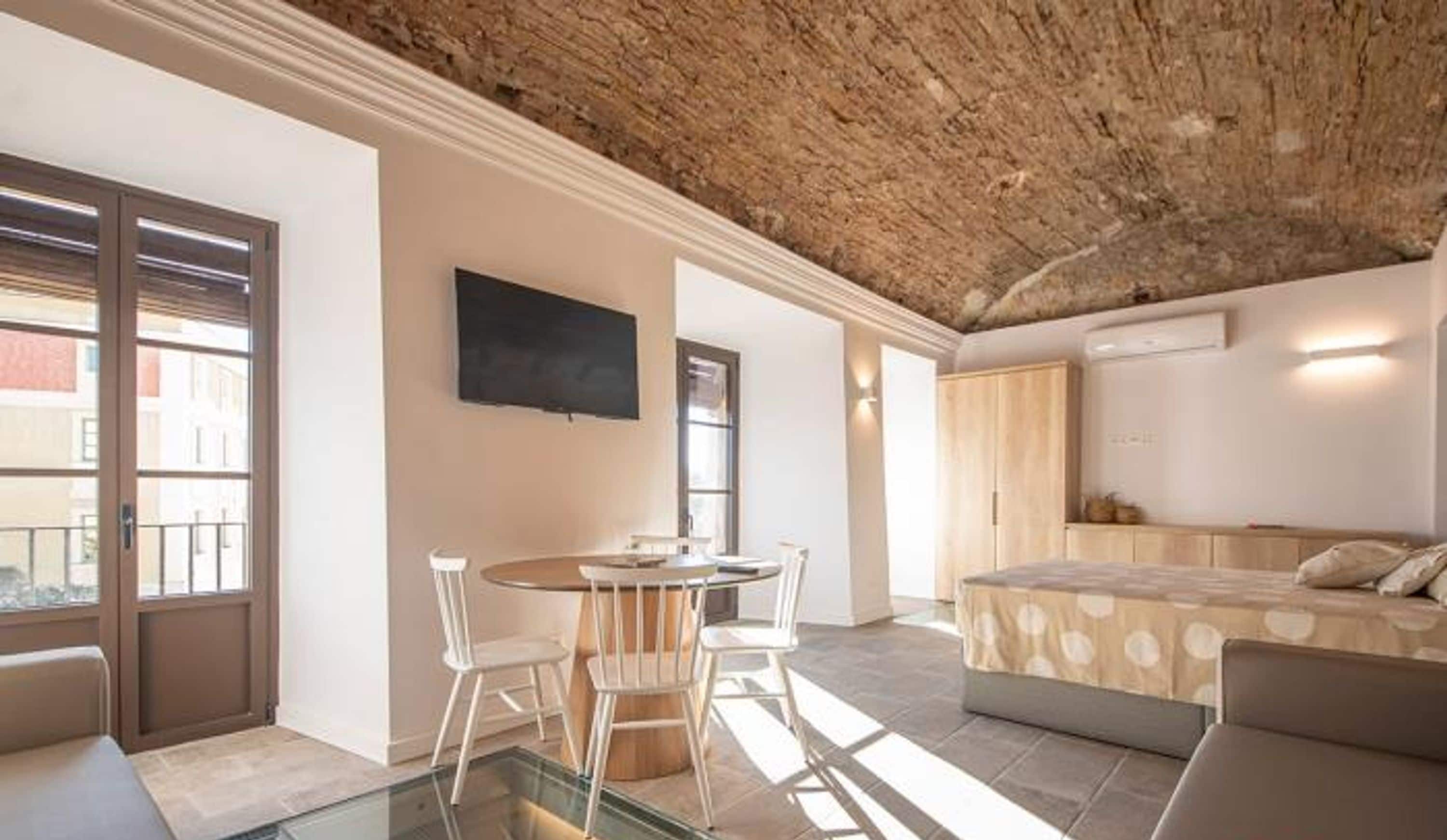 Een modern appartement met een glazen salontafel, een bed met een beddegoed met patroon, een eethoek met stoelen, en een tv aan de muur.