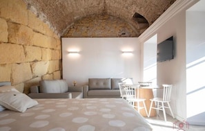 Une chambre avec un mur en pierre, un lit recouvert d’une couvre-lit à pois, un canapé, une petite table avec des chaises et un téléviseur fixé au mur.