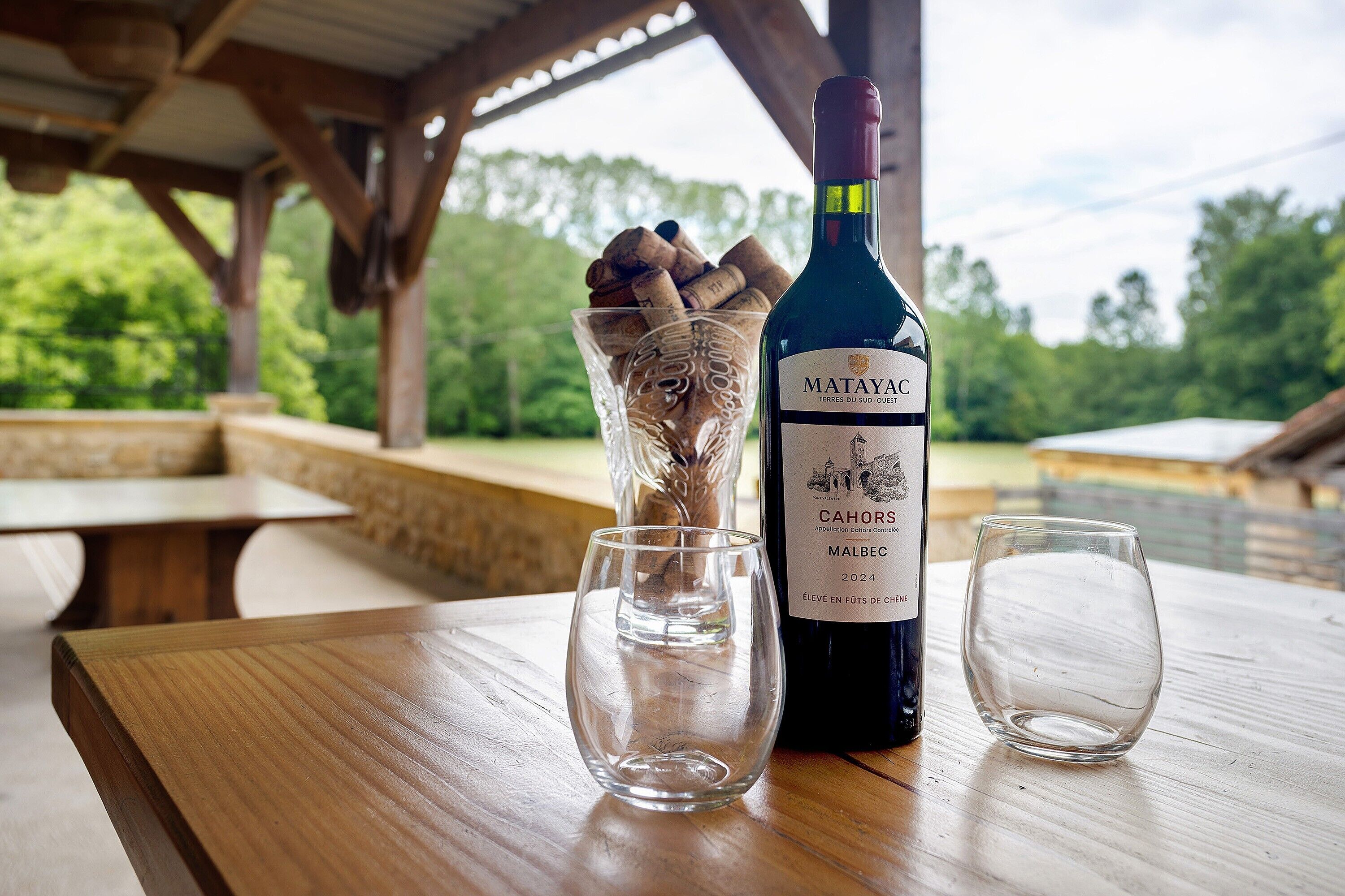 A bottle of Matayac Cahors Malbec 2024 wine, a glass, and a bowl of cinnamon sticks on a wooden table.