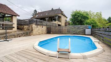 Una piscina con cubierta de madera, un muro de piedra y una casa con techo de tejas.