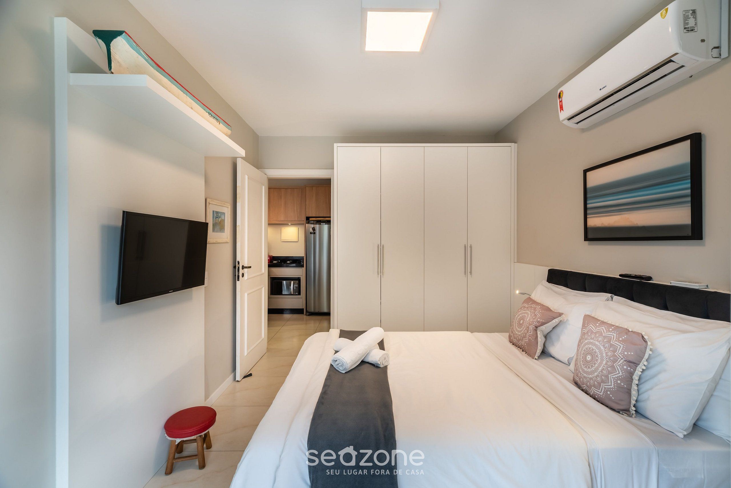 A modern hotel room with a bed, a flat-screen TV, a wardrobe, and a kitchenette in the background.