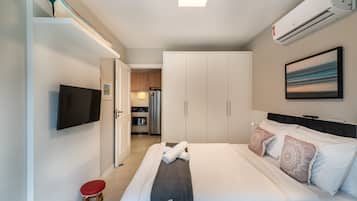 A modern hotel room with a bed, a flat-screen TV, a wardrobe, and a kitchenette in the background.