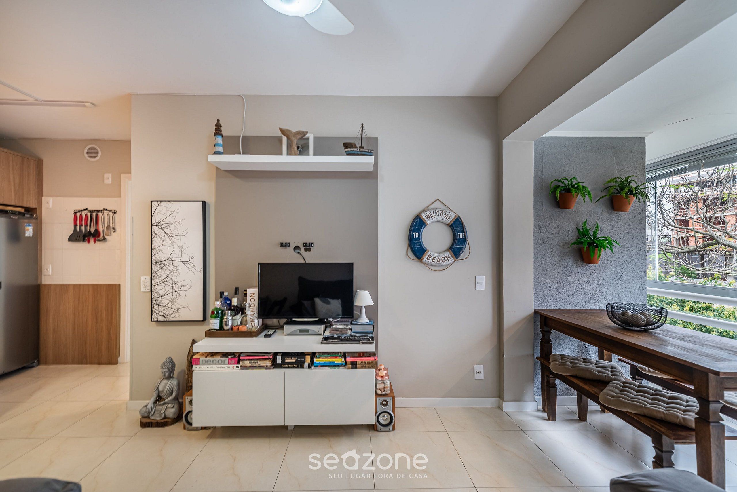 A modern living room with a TV, books, and decorative items on a white console table. There is a wooden bench and a dining table with potted plants.