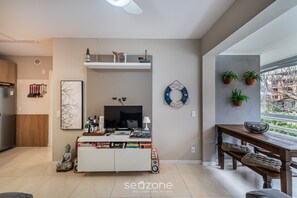 A modern living room with a TV, books, and decorative items on a white console table. There is a wooden bench and a dining table with potted plants.