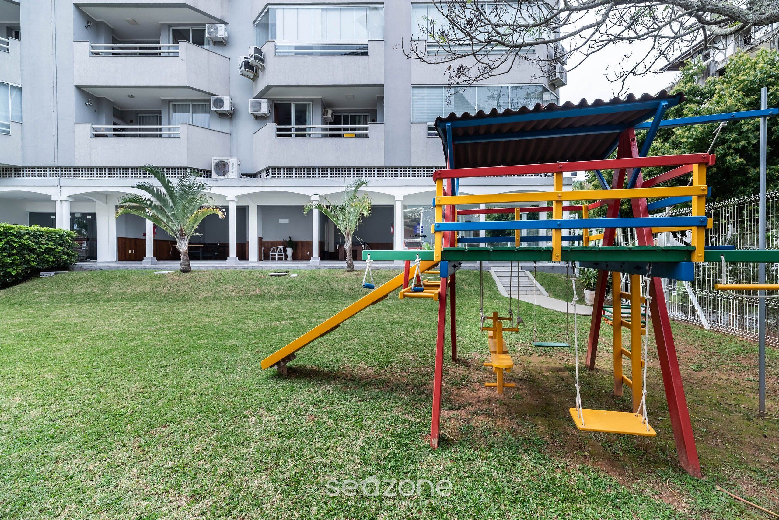 A playground with a slide and swings, surrounded by a grassy area and a building with balconies and air conditioning units.