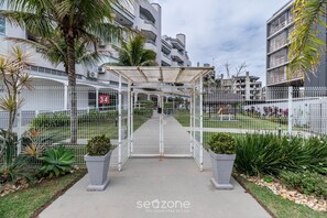 Una entrada con portón a un complejo residencial con un corredor cubierto, palmeras y una cerca blanca.