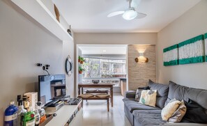 Una sala de estar moderna con un sofá gris, una mesa de centro de madera y un televisor de pantalla plana. Hay un ventilador de techo, una lámpara fijada a la pared y vistas al exterior a través de ventanas amplias.