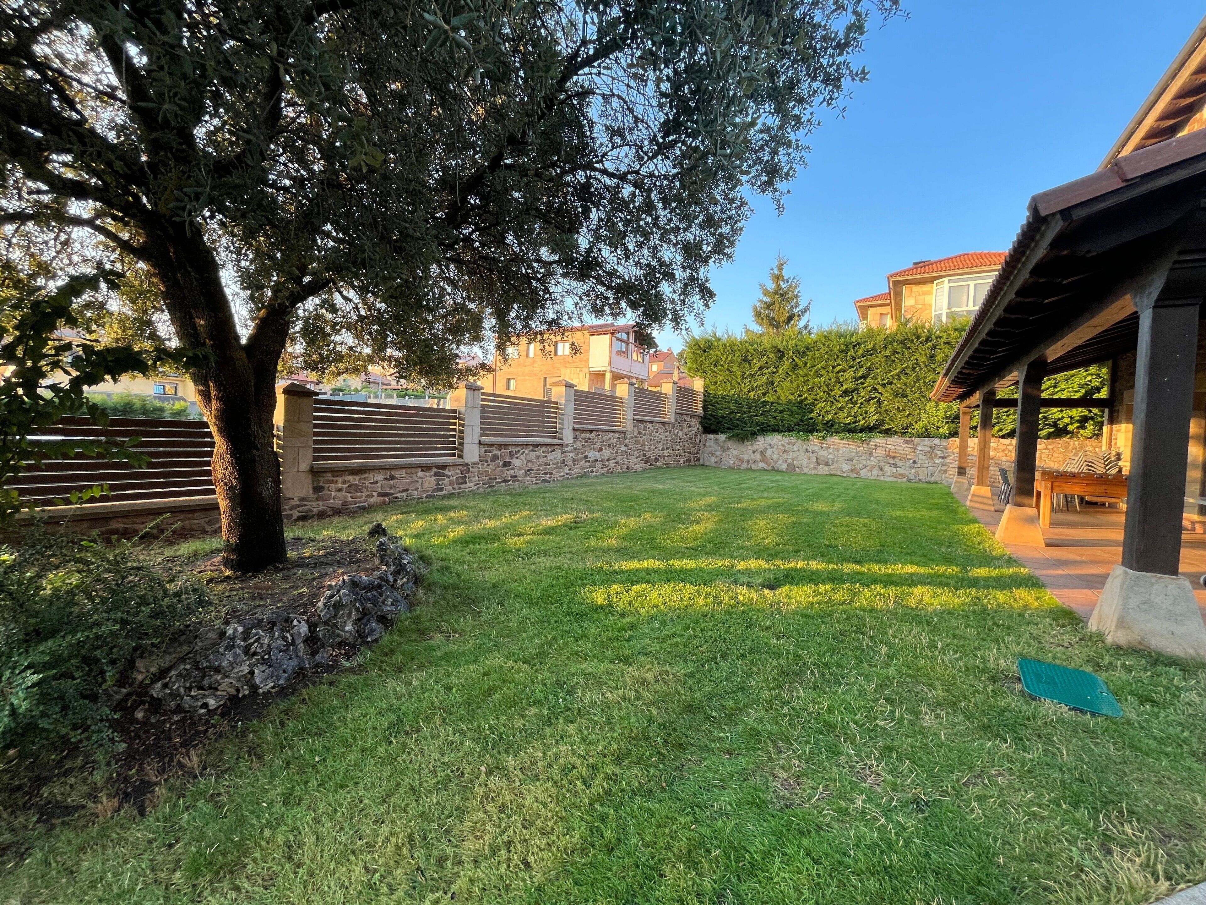 Un patio trasero con césped cuidado, un muro de piedra, una valla de madera y una estructura de pérgola.