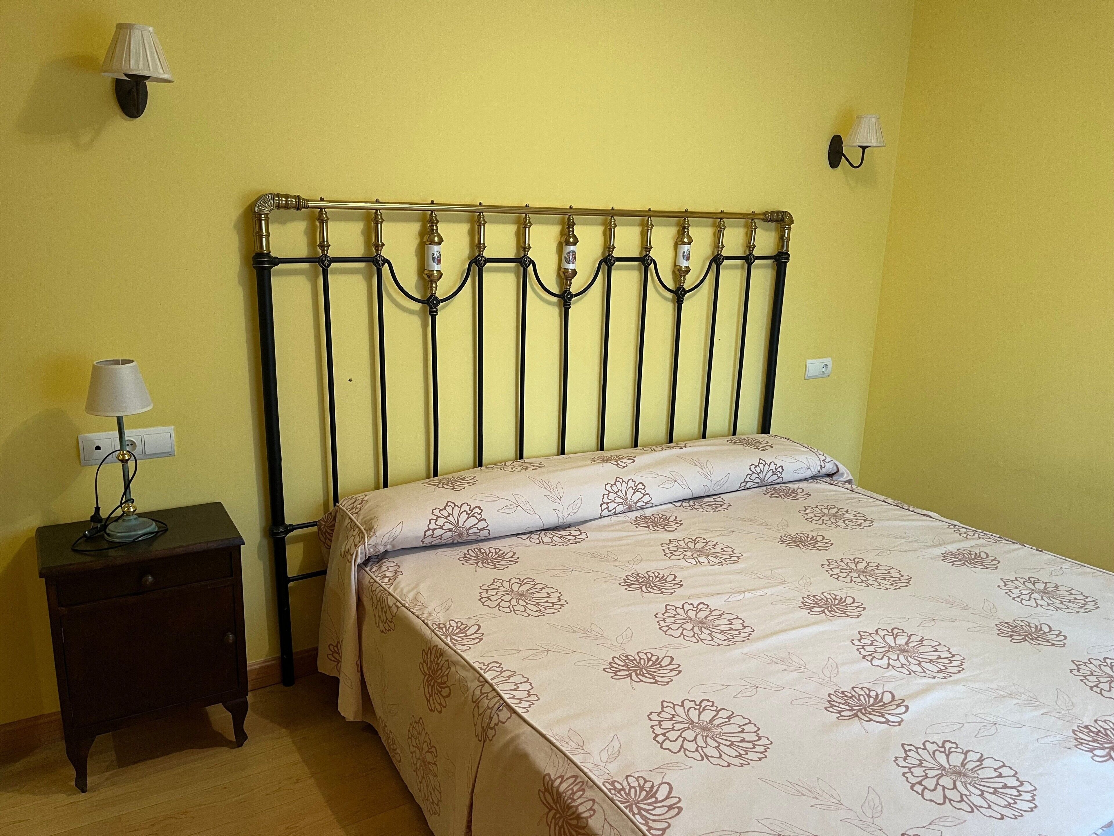Una cama con colcha floral, una mesita de noche con lámpara, y un aplique de pared.