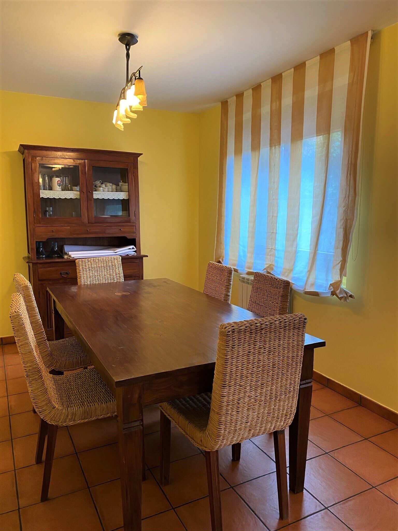 Een eethoek met een houten tafel en rieten stoelen, een kast met glazen deuren en een raam met gestreepte gordijnen.