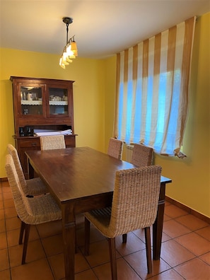 A dining area with a wooden table and wicker chairs, a cabinet with glass doors, and a window with striped curtains.